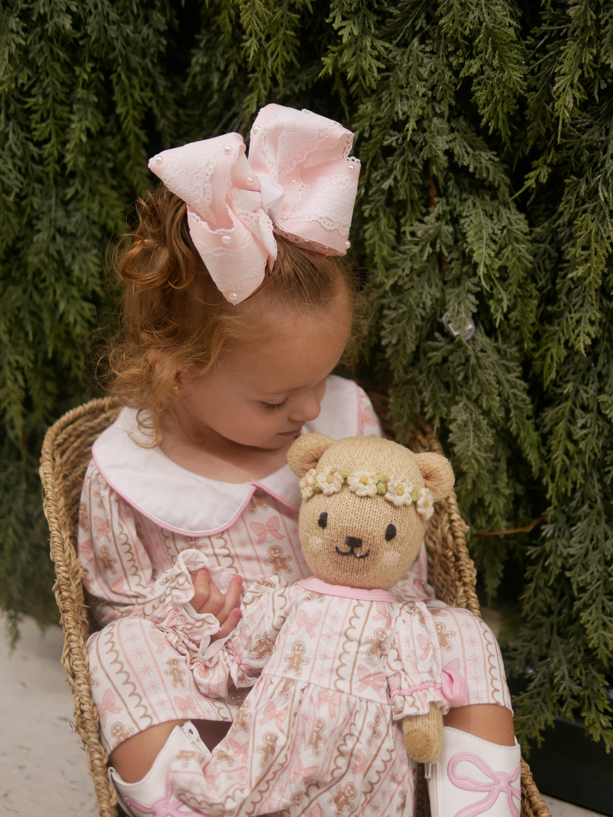 A young child wearing a pink and white patterned dress with a large matching pink bow in their hair, sitting in a wicker chair and holding a teddy bear dressed in a matching pink outfit against a background of green foliage.
