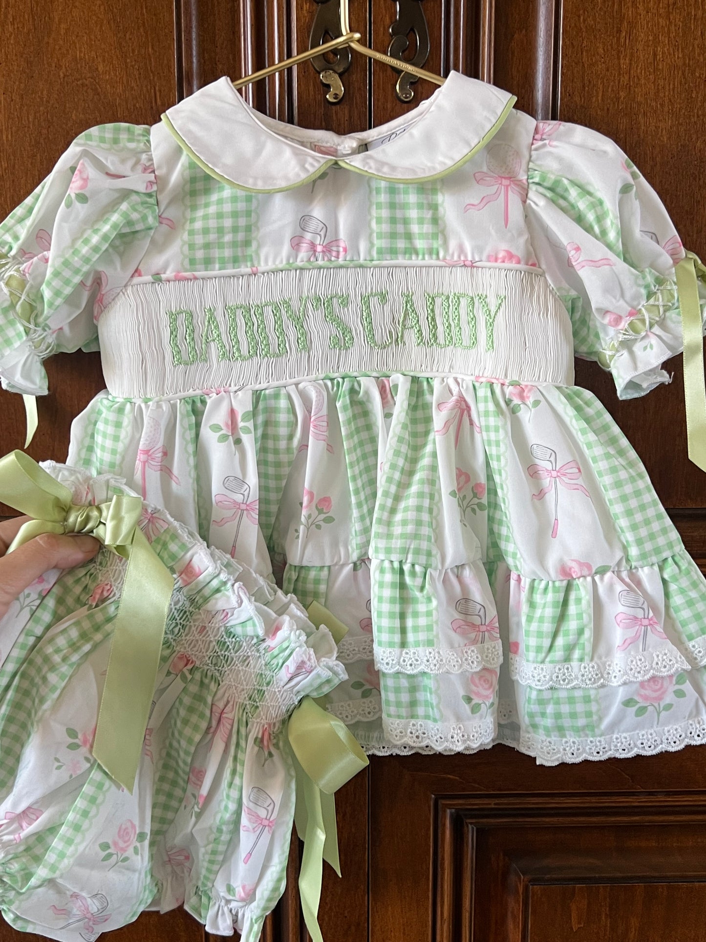 A baby girl's smocked dress with a green and white gingham pattern, pink bow and golf motifs, and 'Daddys Caddy' smocked on the chest. A hand is holding a matching smocked bonnet with a green ribbon, set against a dark wood background.
