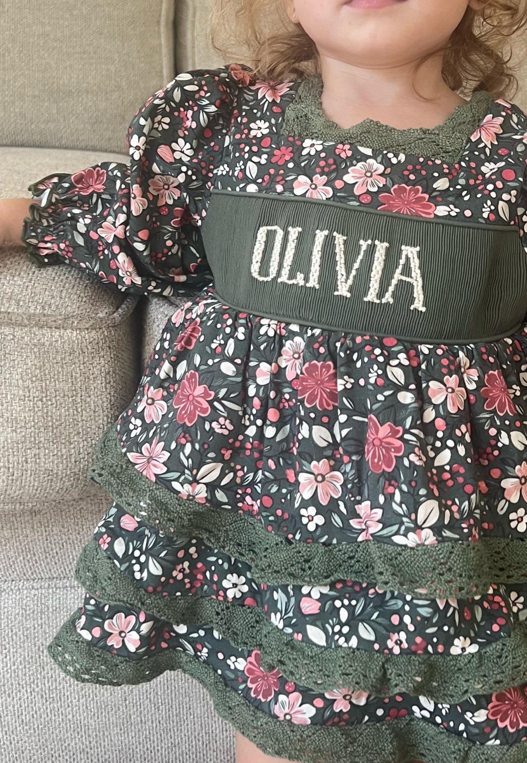 A close-up of a young child wearing a dark green tiered dress with a pink and white floral print, featuring a wide green waistband with the name 'OLIVIA' in white lettering.