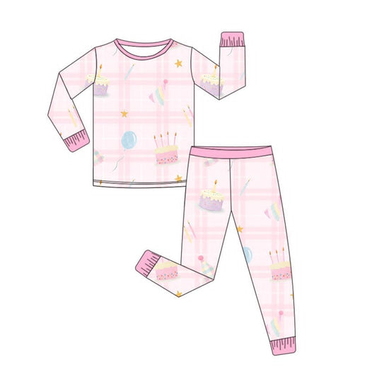 A two-piece children's pajama set shown as a flat lay illustration, featuring a long-sleeve top and matching pants. The set has a light pink and white plaid pattern with a birthday-themed print of cakes, candles, stars, and balloons, accented with pink trim.