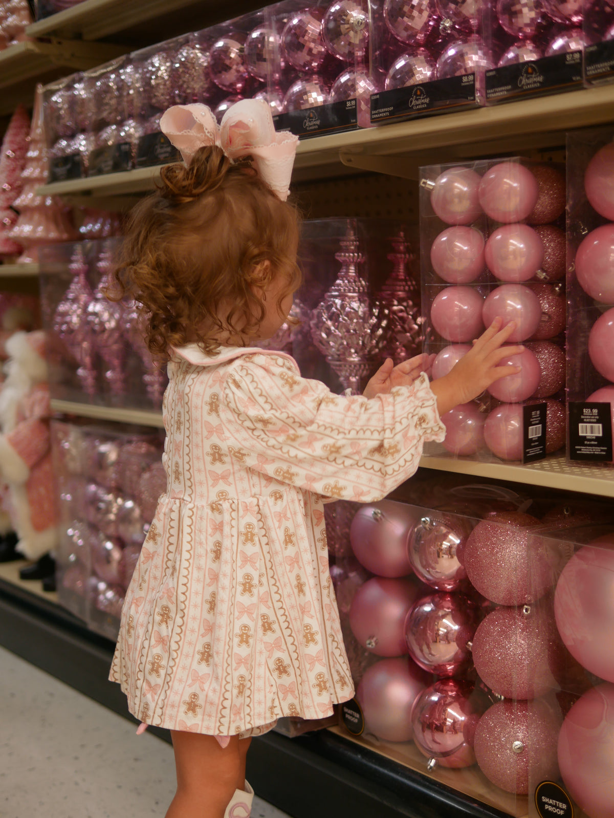 A young child wearing a pink and white patterned dress and a large pink bow in her hair reaches for a pink Christmas ornament on a store shelf.