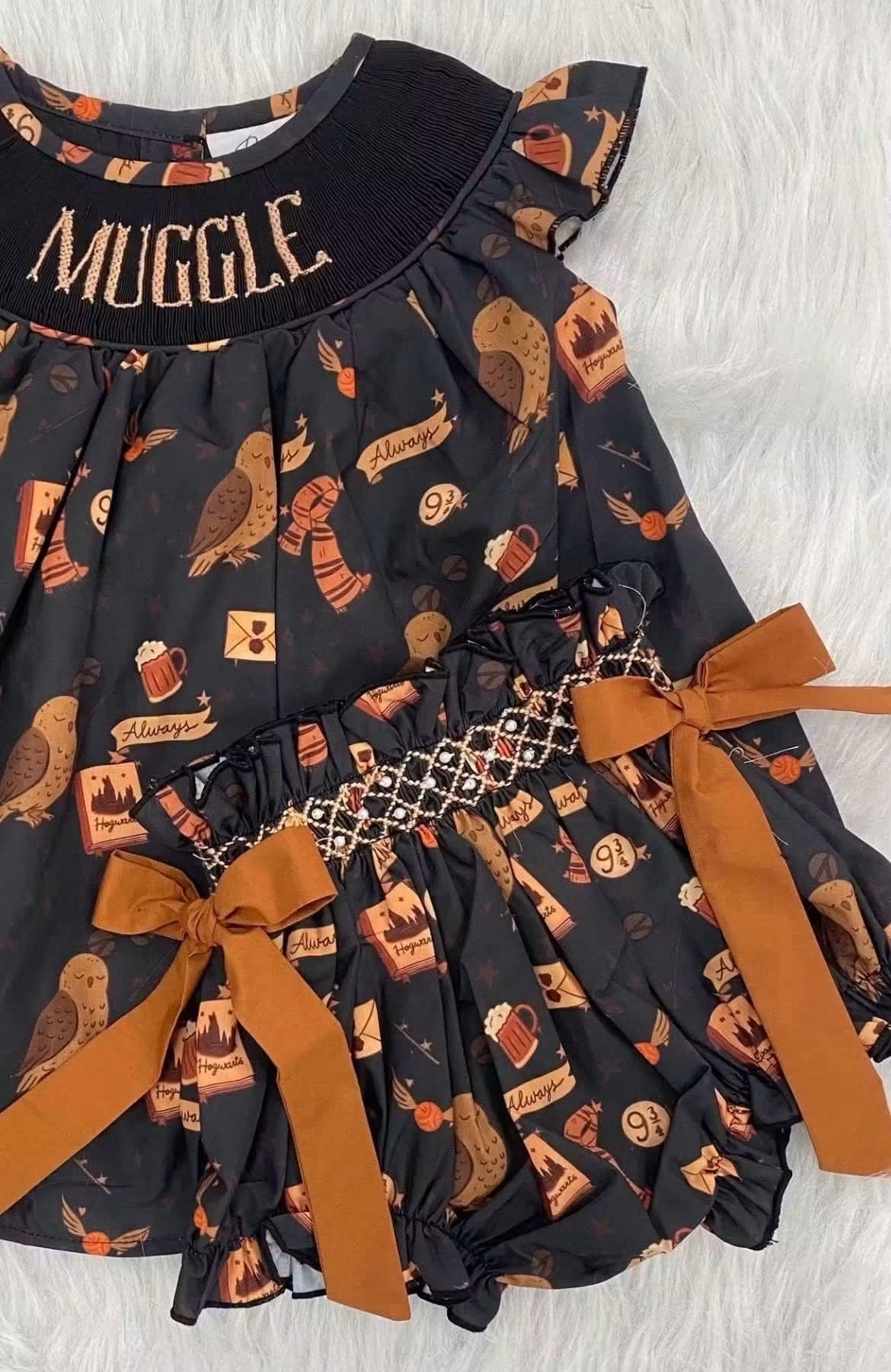 A toddler girl's dress featuring a black base with an all-over HP Muggle motif, including owls, cauldron, and potion bottles. The dress has a black ruffled collar with 'MUGGLE' embroidered in gold, a smocked waist, and two large brown ribbon bows on the skirt.