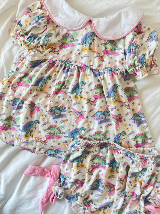 A baby girl's two-piece romper set featuring a colorful dinosaur print on a cream-colored fabric, complete with a white Peter Pan collar trimmed in pink and matching ruffled bloomers with a pink ribbon tie, laid flat on a white sheet.