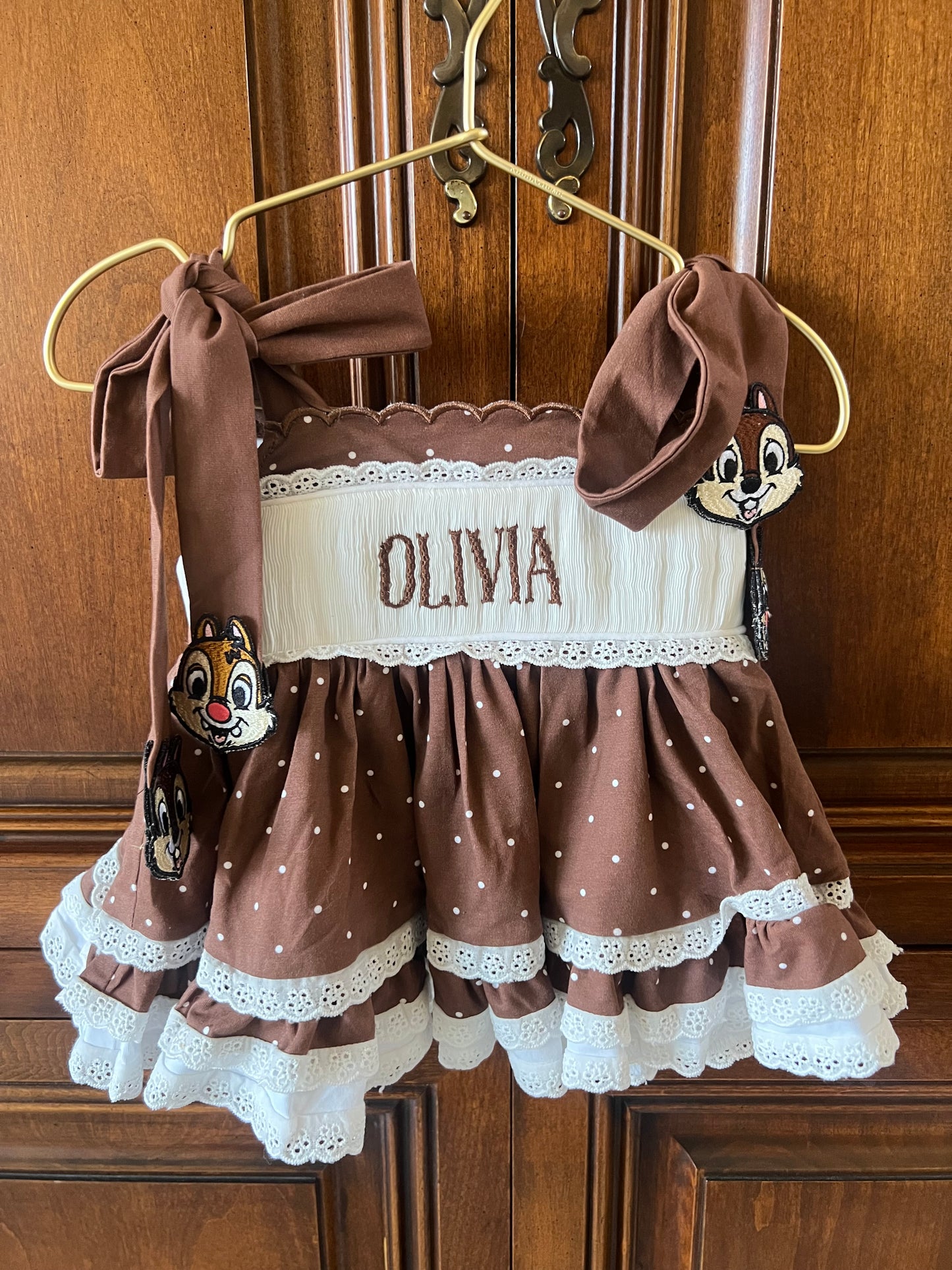 A brown and white polka dot baby dress with 'OLIVIA' embroidered on the front, hanging on a gold hanger against a wooden door. The dress features a white lace-trimmed bodice, brown polka dot skirt, and matching brown ribbon bows on the shoulders.