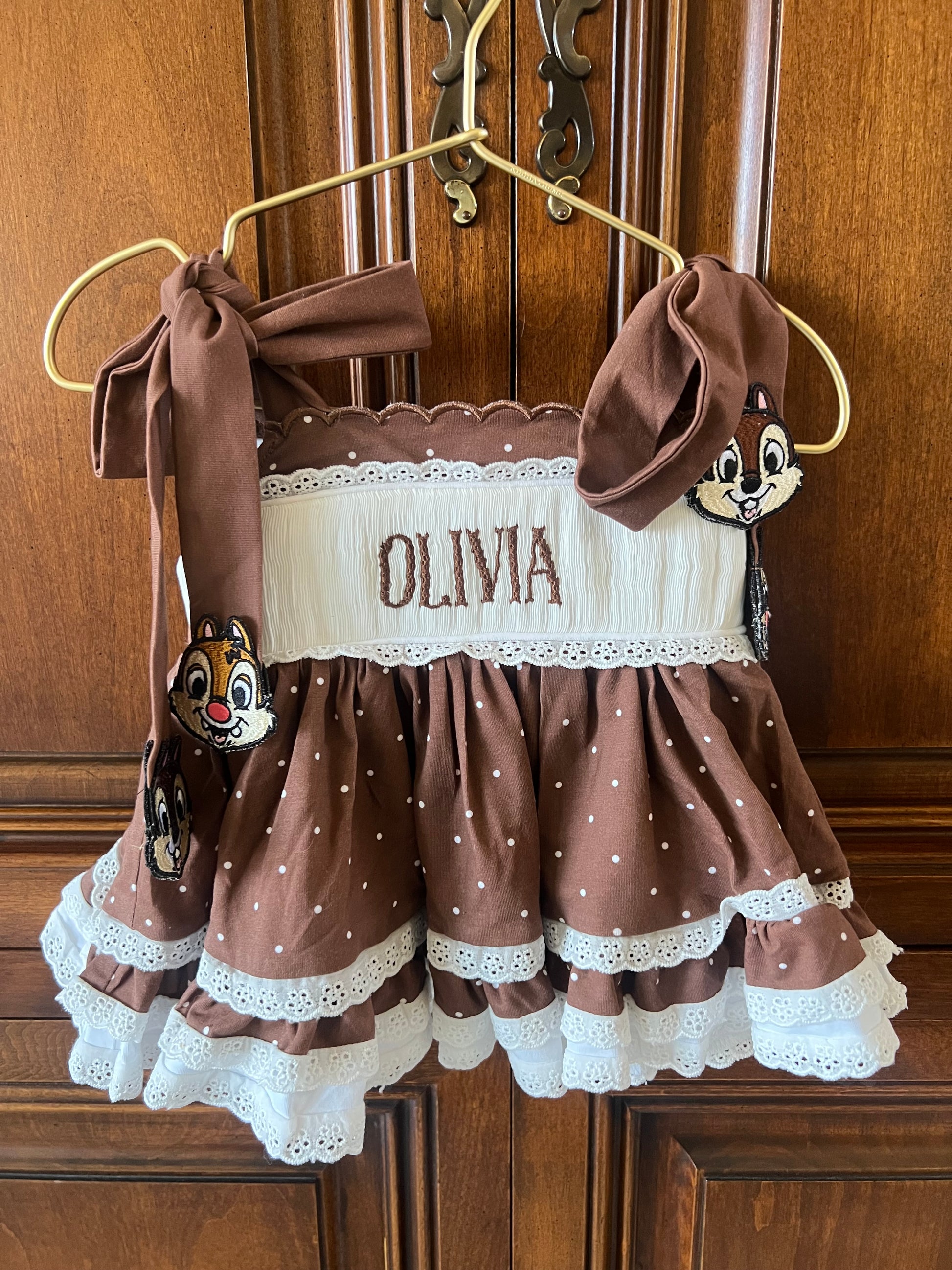 A brown and white polka dot baby dress with 'OLIVIA' embroidered on the front, hanging on a gold hanger against a wooden door. The dress features a white lace-trimmed bodice, brown polka dot skirt, and matching brown ribbon bows on the shoulders.