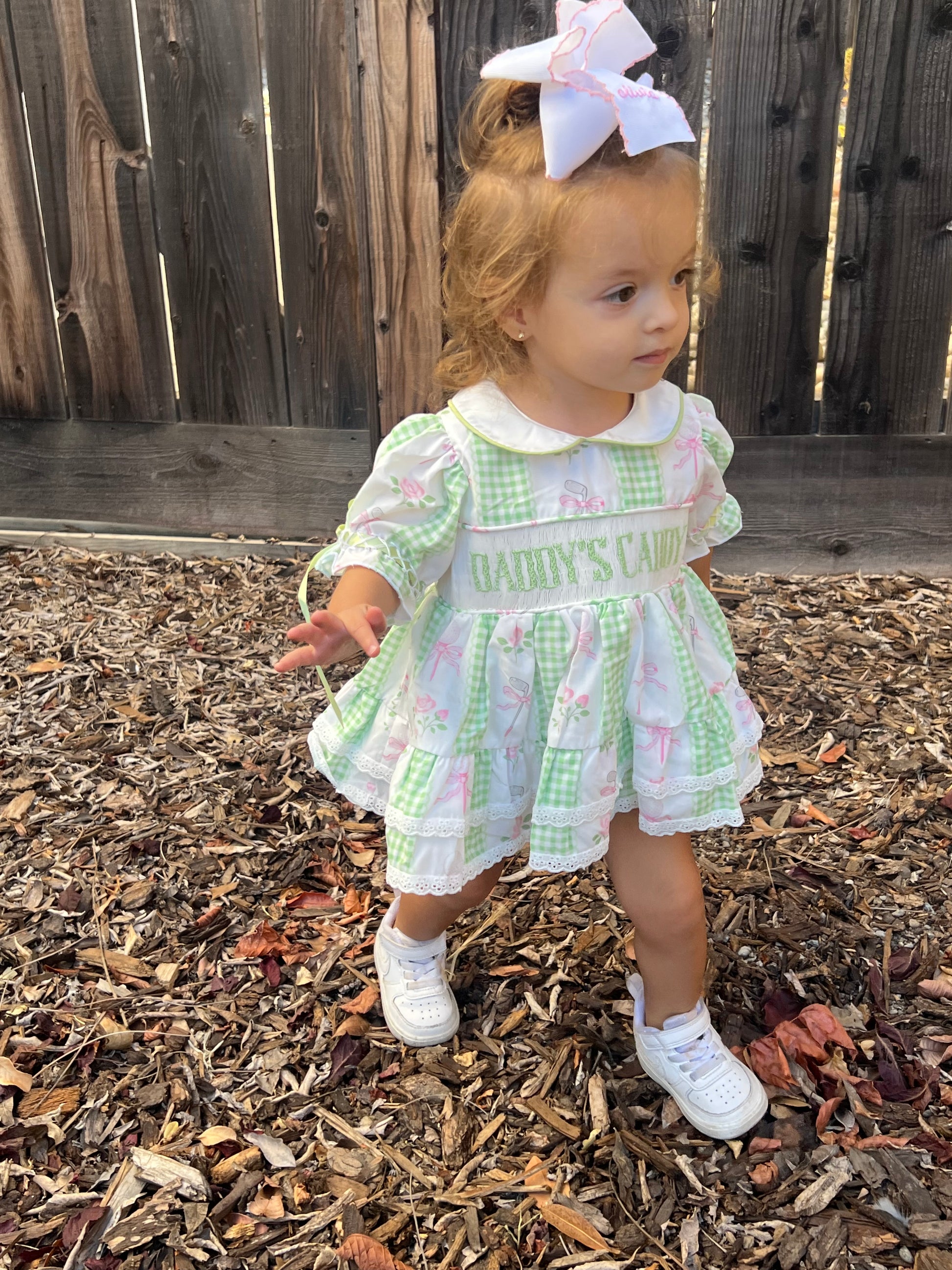 A toddler girl wearing a green and white gingham dress with golf prints and 'daddys caddy' on the chest, standing outdoors on a wood mulch surface in front of a wooden fence.