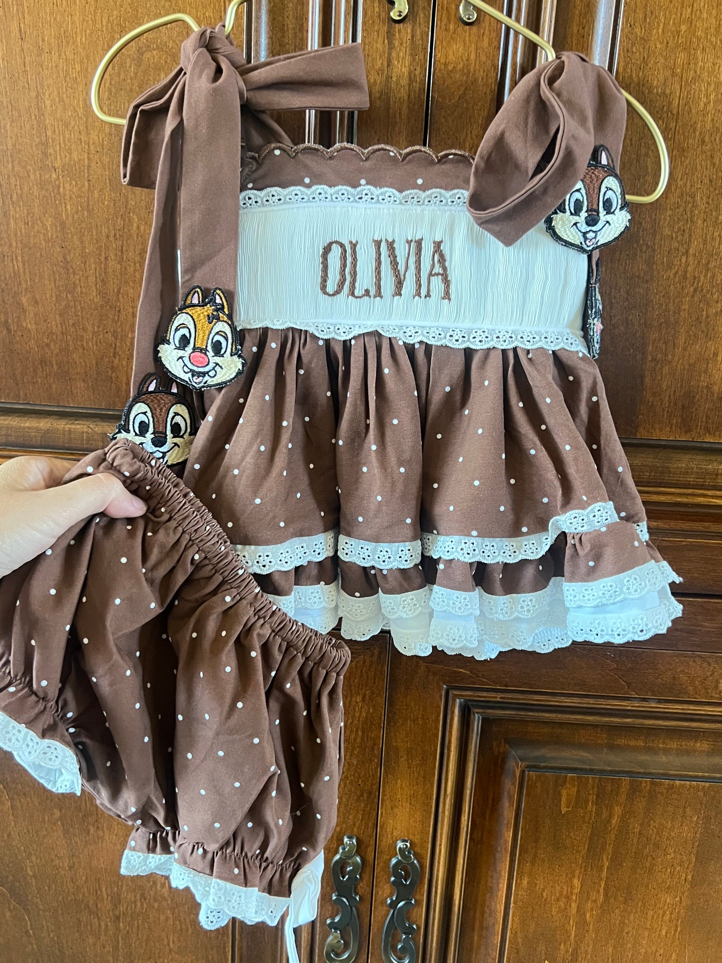 A brown and white polka dot baby girl's two-piece outfit set consisting of a ruffled dress and matching bloomers, featuring a white embroidered name panel that reads 'OLIVIA' and character face patches, hanging on a wooden door.