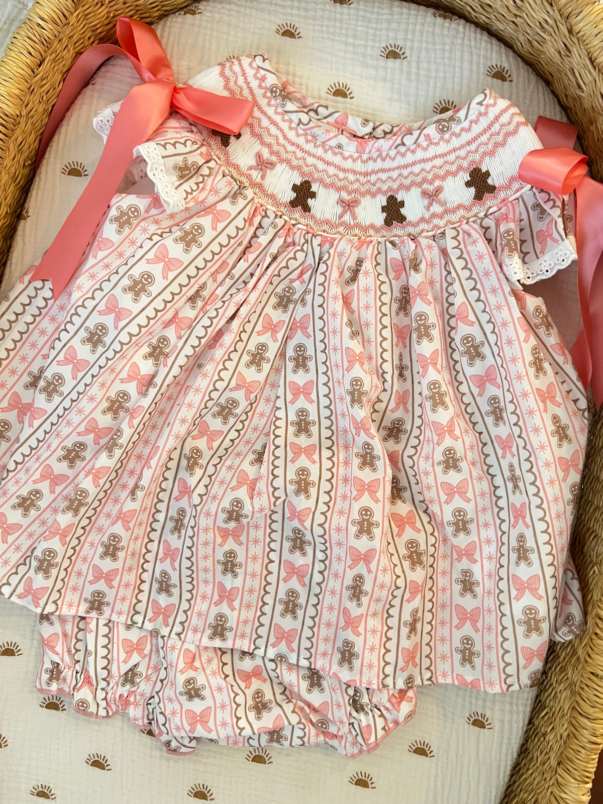 A baby girl's dress and matching bloomers set in a woven bassinet. The outfit features a white base with vertical stripes of brown gingerbread men, pink bows, and a pink and white patterned yoke with embroidered brown gingerbread men.