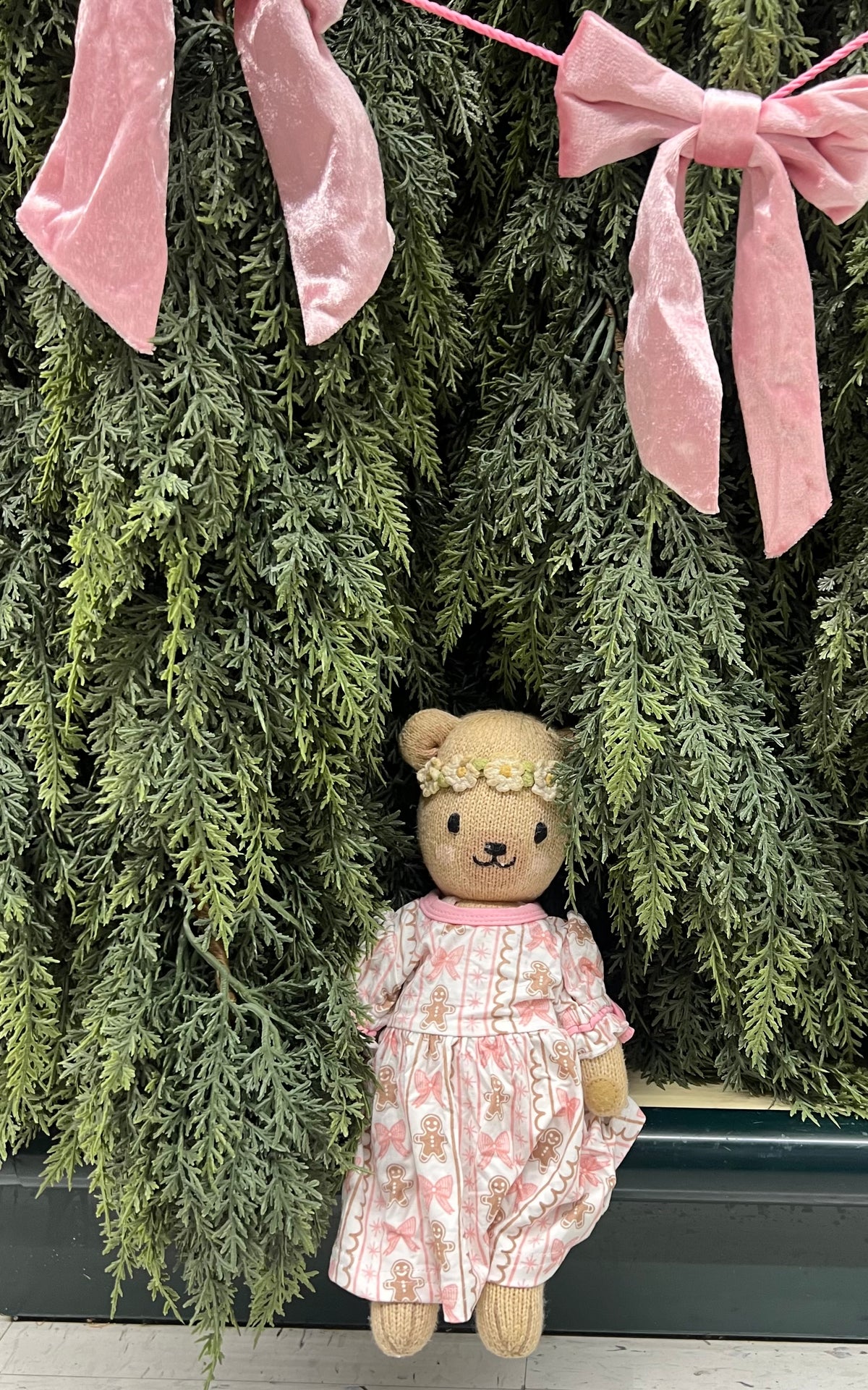 A teddy bear doll wearing a pink floral dress and a small flower crown, posed against a green evergreen garland with pink velvet bows.