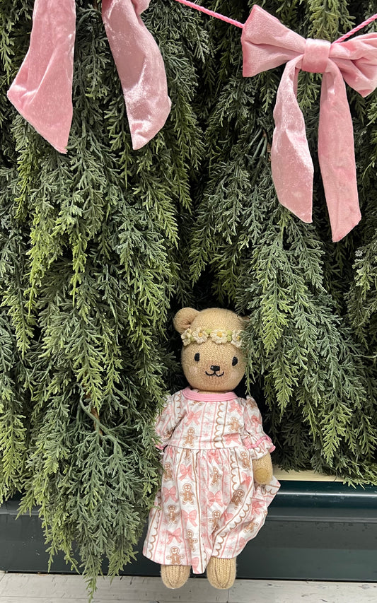 A teddy bear doll wearing a pink floral dress and a small flower crown, posed against a green evergreen garland with pink velvet bows.