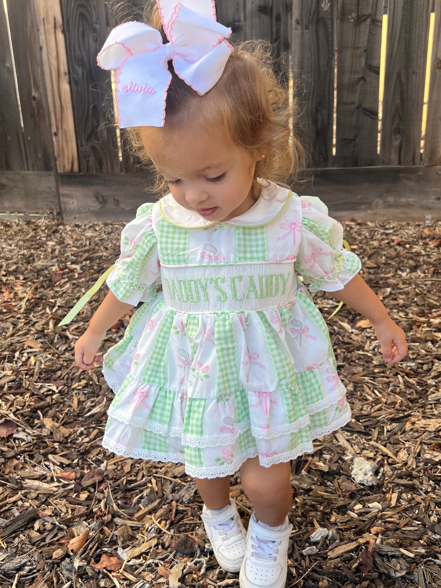 A toddler girl wearing a green and white gingham dress with pink bunny prints, white lace trim, and 'Daddys caddy embroidered on the chest, standing outdoors on a wood mulch surface with a wooden fence behind her.