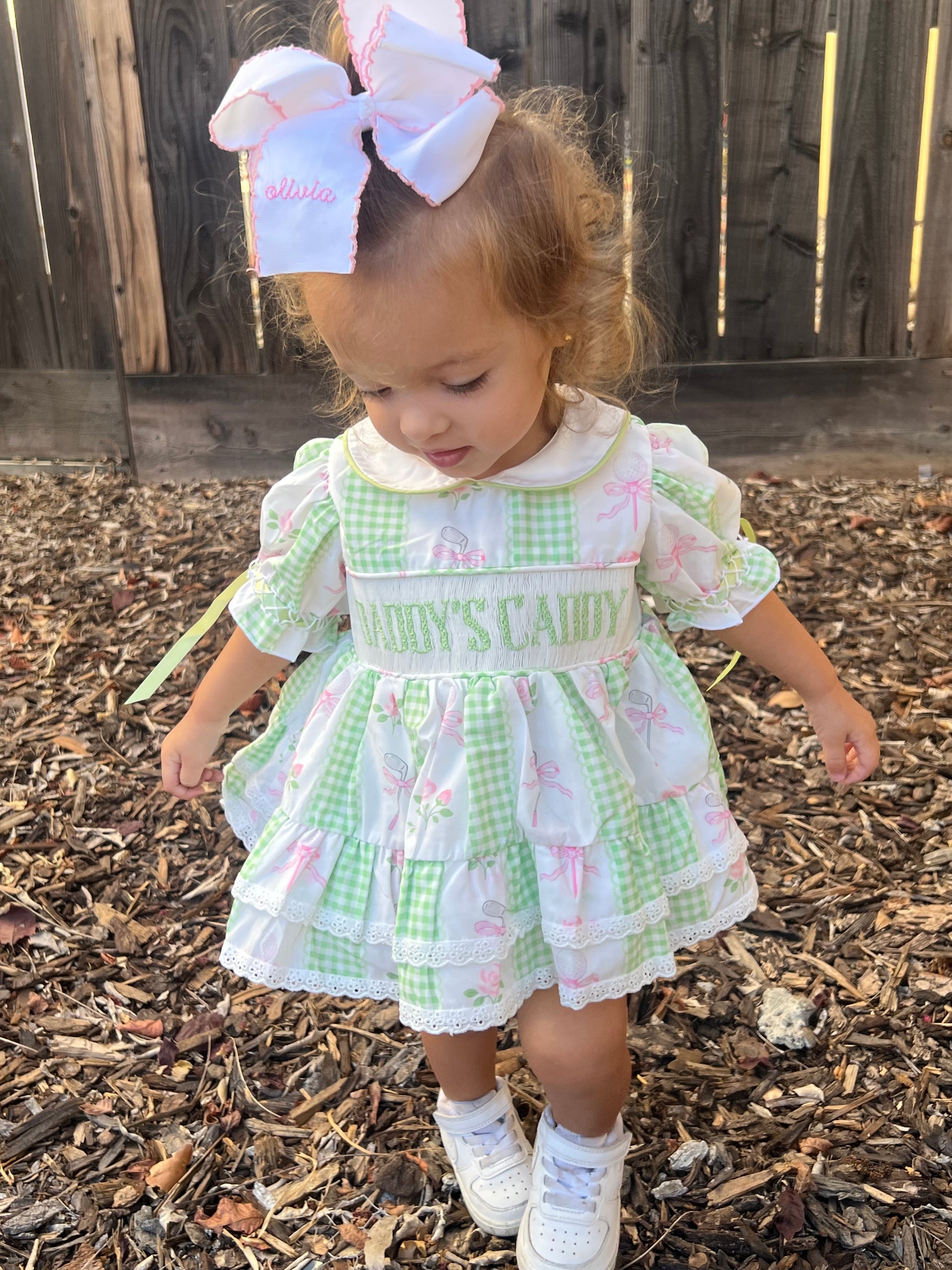 A toddler girl wearing a green and white gingham dress with pink bunny prints, white lace trim, and 'Daddys caddy embroidered on the chest, standing outdoors on a wood mulch surface with a wooden fence behind her.