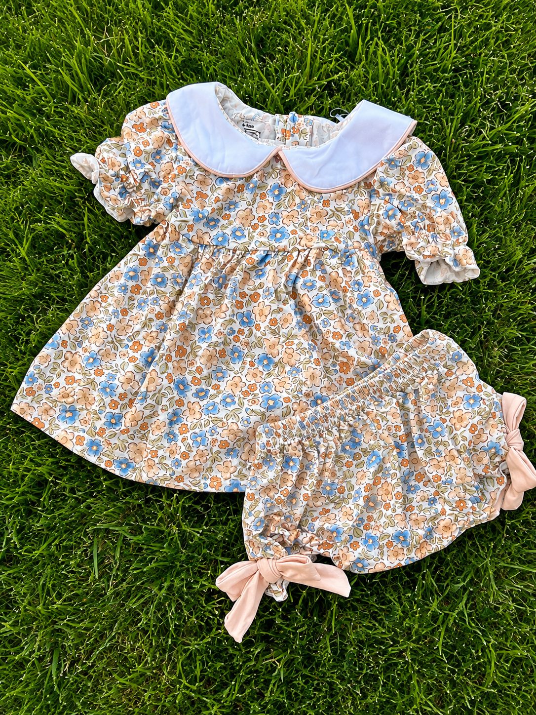 A baby girl's floral dress and matching bloomers laid flat on green grass. The dress has a cream base with a blue and orange floral print, a white Peter Pan collar, and short puff sleeves.