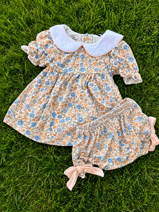A baby girl's floral dress and matching bloomers laid flat on green grass. The dress has a cream base with a blue and orange floral print, a white Peter Pan collar, and short puff sleeves.