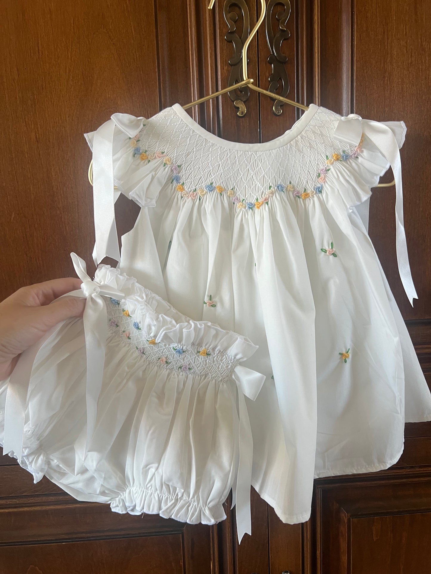 A white baby girl's two-piece outfit consisting of a smocked blouse with ruffled sleeves and matching smocked bloomers, held up by a hand in front of a dark wood door.