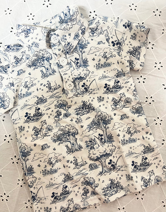 A short-sleeve button-front shirt with a blue and white toile de Jouy pattern featuring Disney characters like Mickey Mouse, laid flat on a white embroidered lace surface.